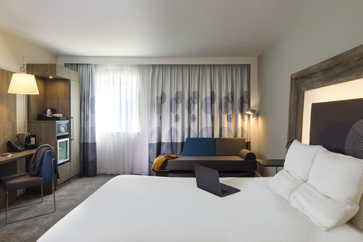 A hotel room with a bed, a desk, a chair, a sofa, a lamp, and a large window with curtains.