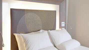 Camera d'albergo con un letto, biancheria bianca, una testiera con motivo geometrico e una lampada da comodino.