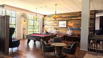 A billiards table in a room with a herringbone floor, wooden walls, and a large window.