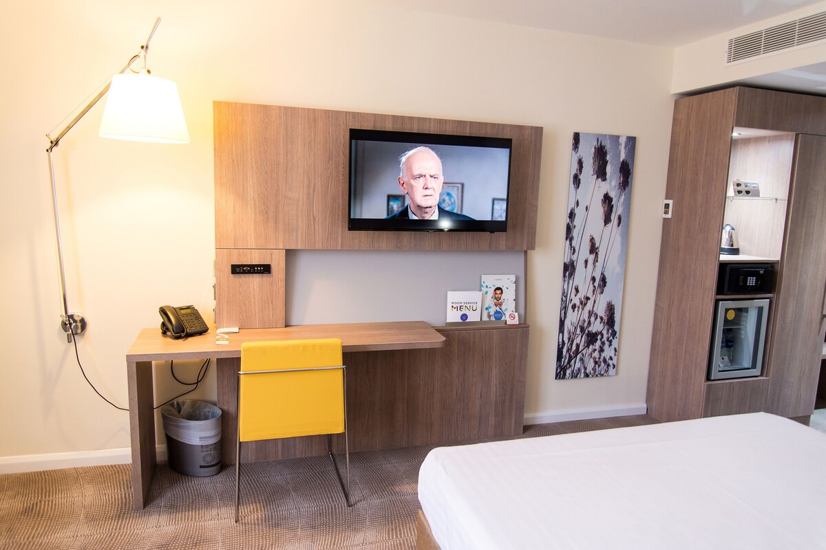 A hotel room with a bed, a desk with a lamp, a television, and a small kitchenette.