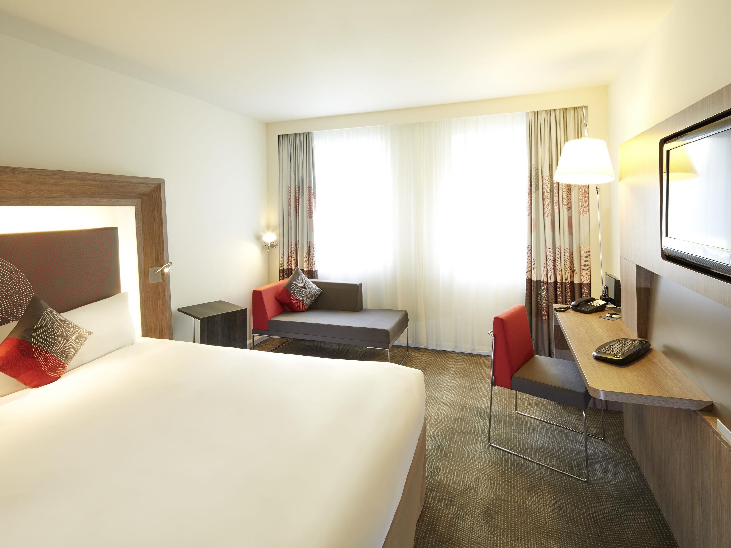 Une chambre d’hôtel avec un lit, un bureau équipé d’un ordinateur, une chaise, un téléviseur et une fenêtre avec des rideaux.
