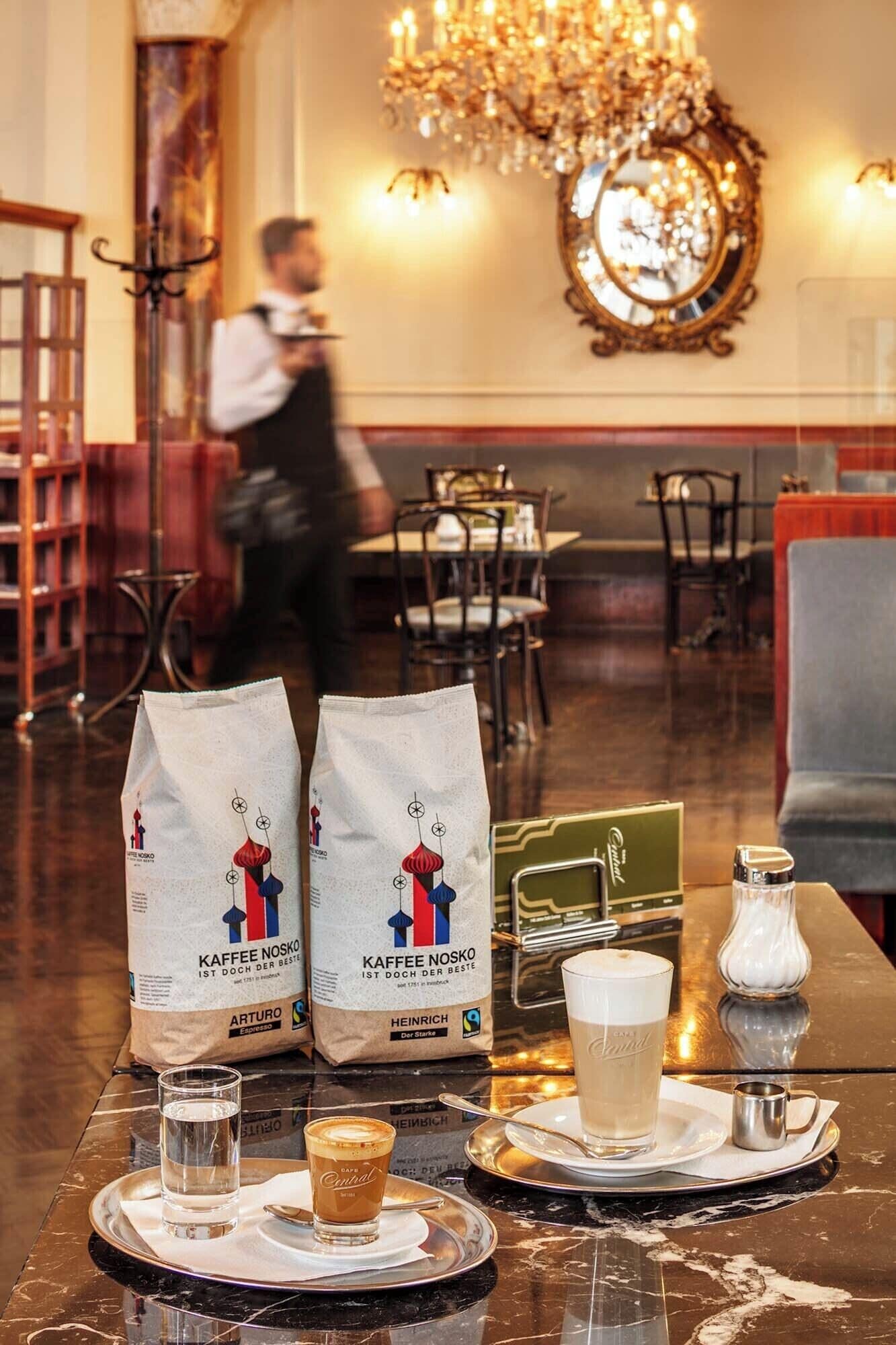 A restaurant setting with a marble table displaying coffee bags, a glass of coffee, and a cup of water.