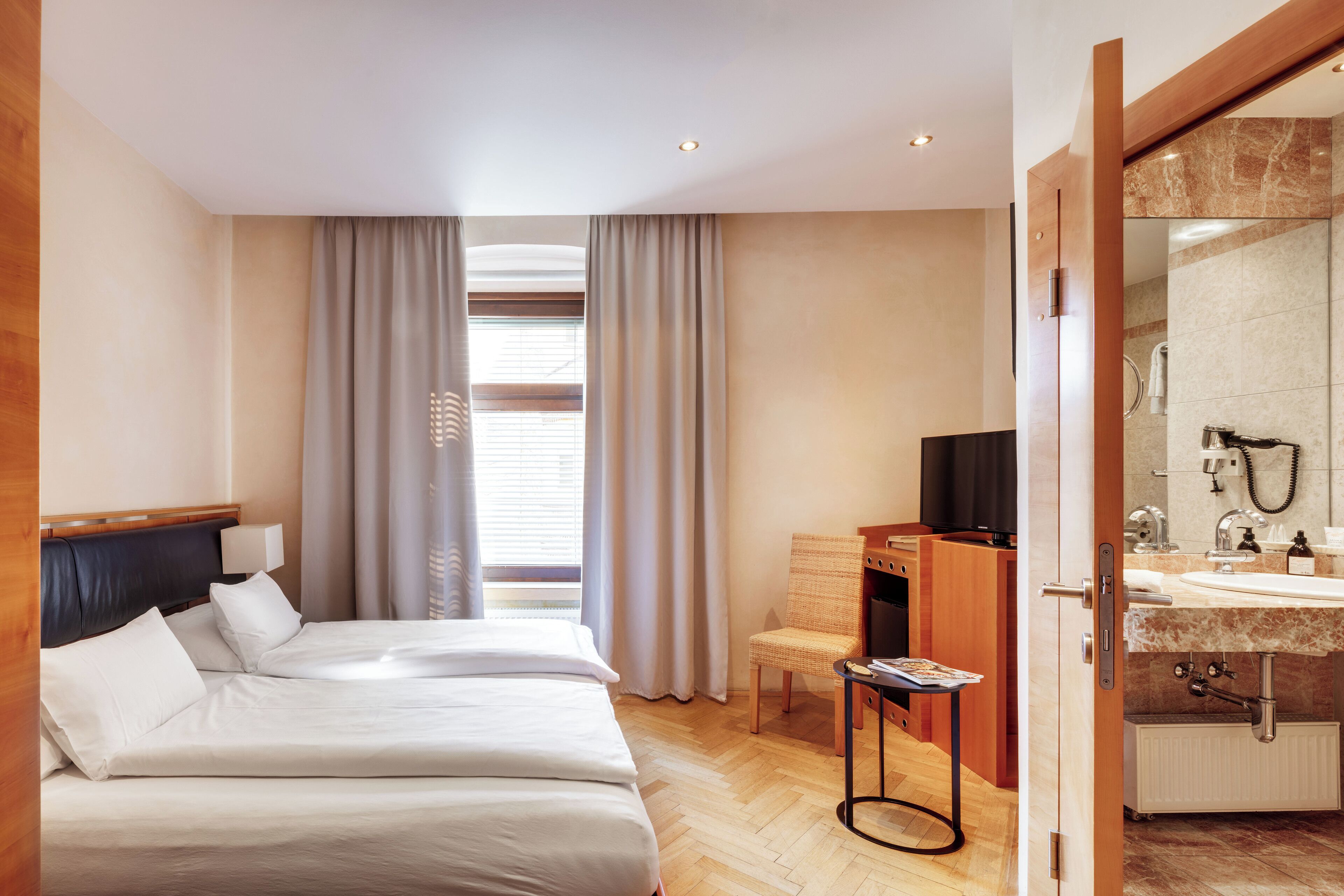 A hotel room with a bed, a desk, a chair, a bathroom with a sink and mirror, and a window with curtains.