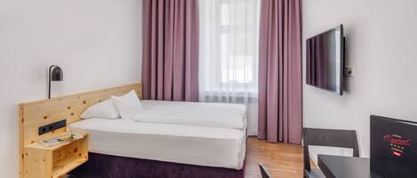 Habitación de hotel con una cama de cabecera de madera, una mesita de noche pequeña de madera, un escritorio negro y un televisor en la pared.