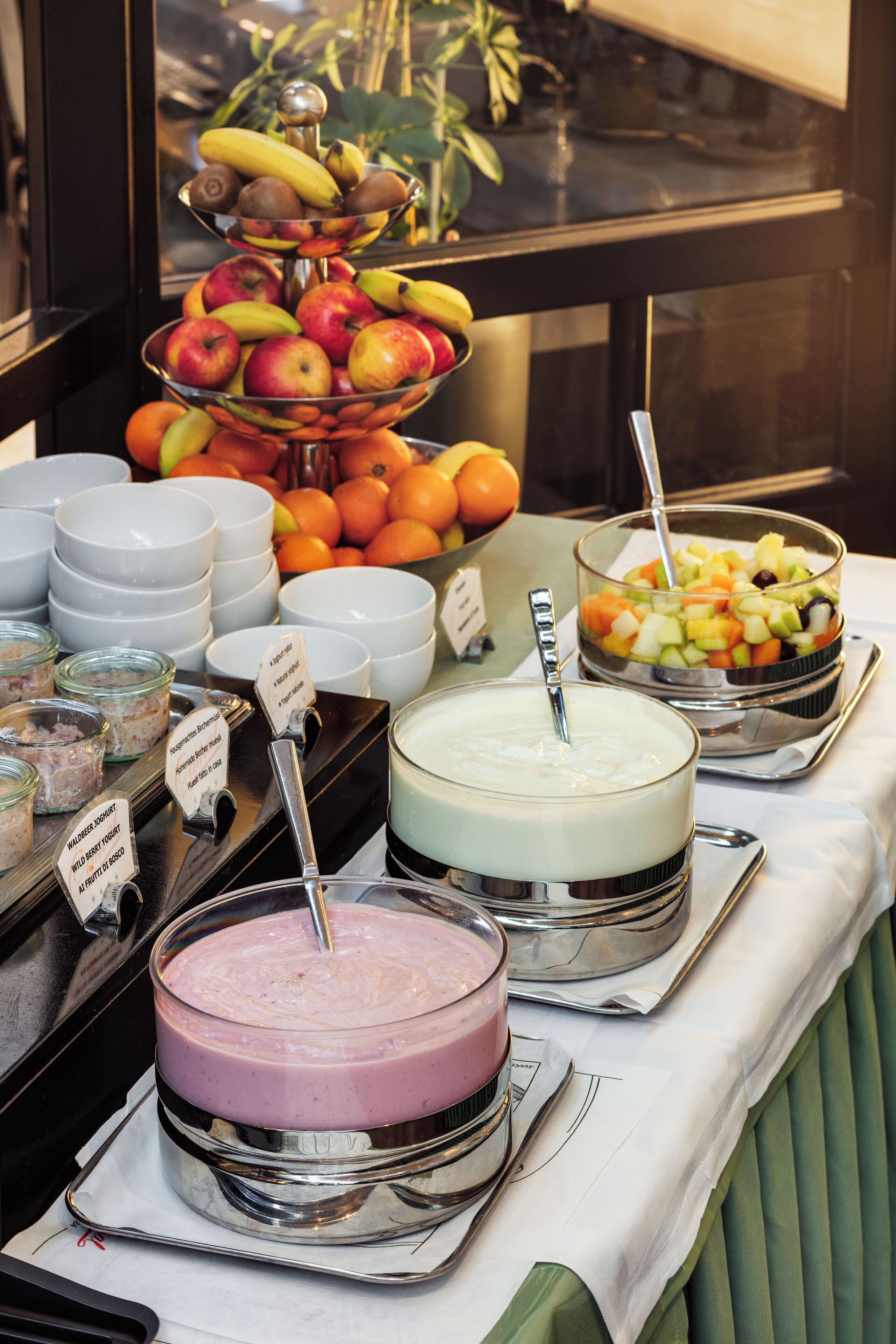Een buffet met diverse gerechten, waaronder een fruitkom in drie niveaus, kommen met yoghurt en een grote taart.