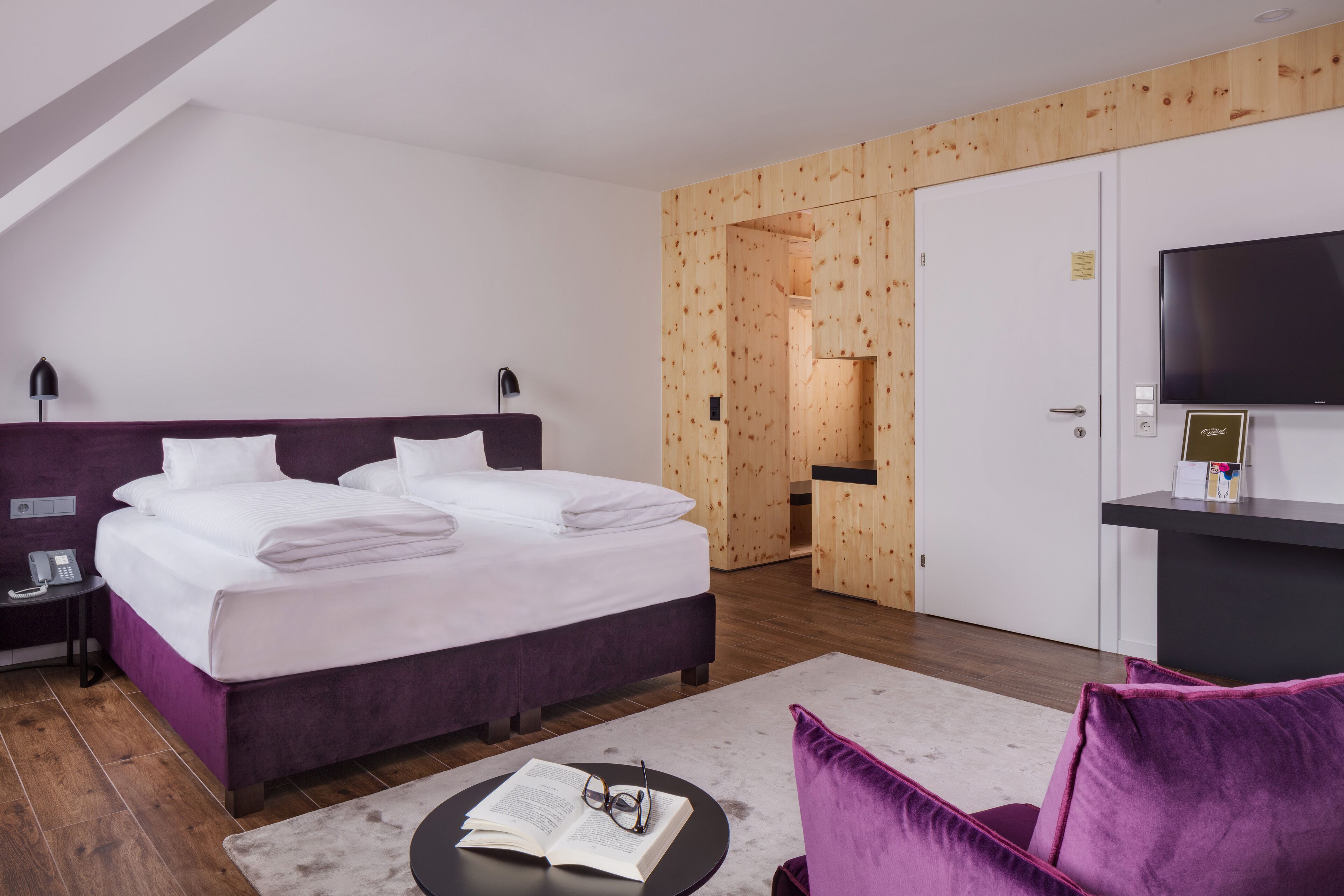 A modern hotel room with a large bed, a desk with a flat-screen TV, a wooden wall, and a small seating area.