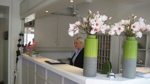 A reception desk with a woman behind it, two green and grey vases with white flowers, and a mirror above the counter. - Hotel S16 (Munich)