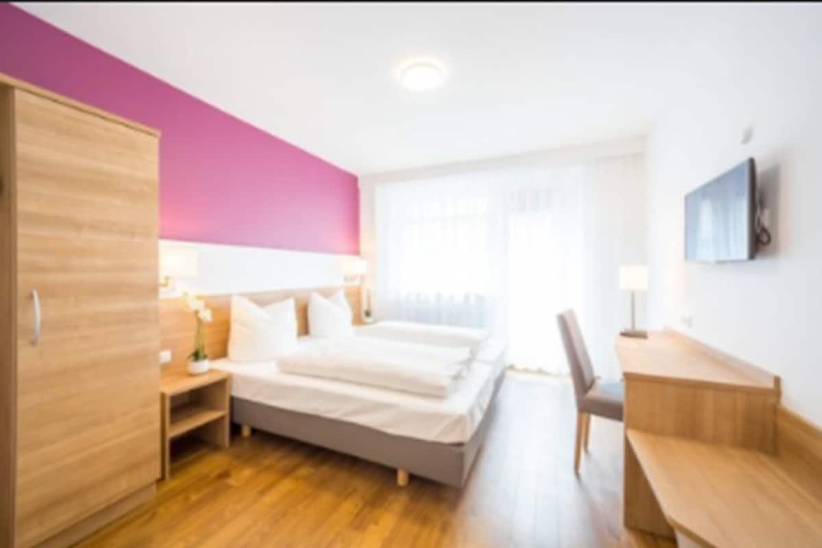 A modern hotel room with a wooden wardrobe, a bed with white linens, a desk, and a chair.