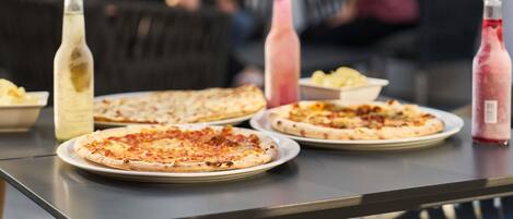 Uma mesa com pizzas e garrafas de bebidas, um sofá, e pessoas ao fundo.