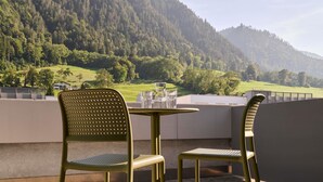 Un balcon avec deux chaises et une table, donnant sur un paysage montagneux et offrant une vue sur une maison et la verdure.