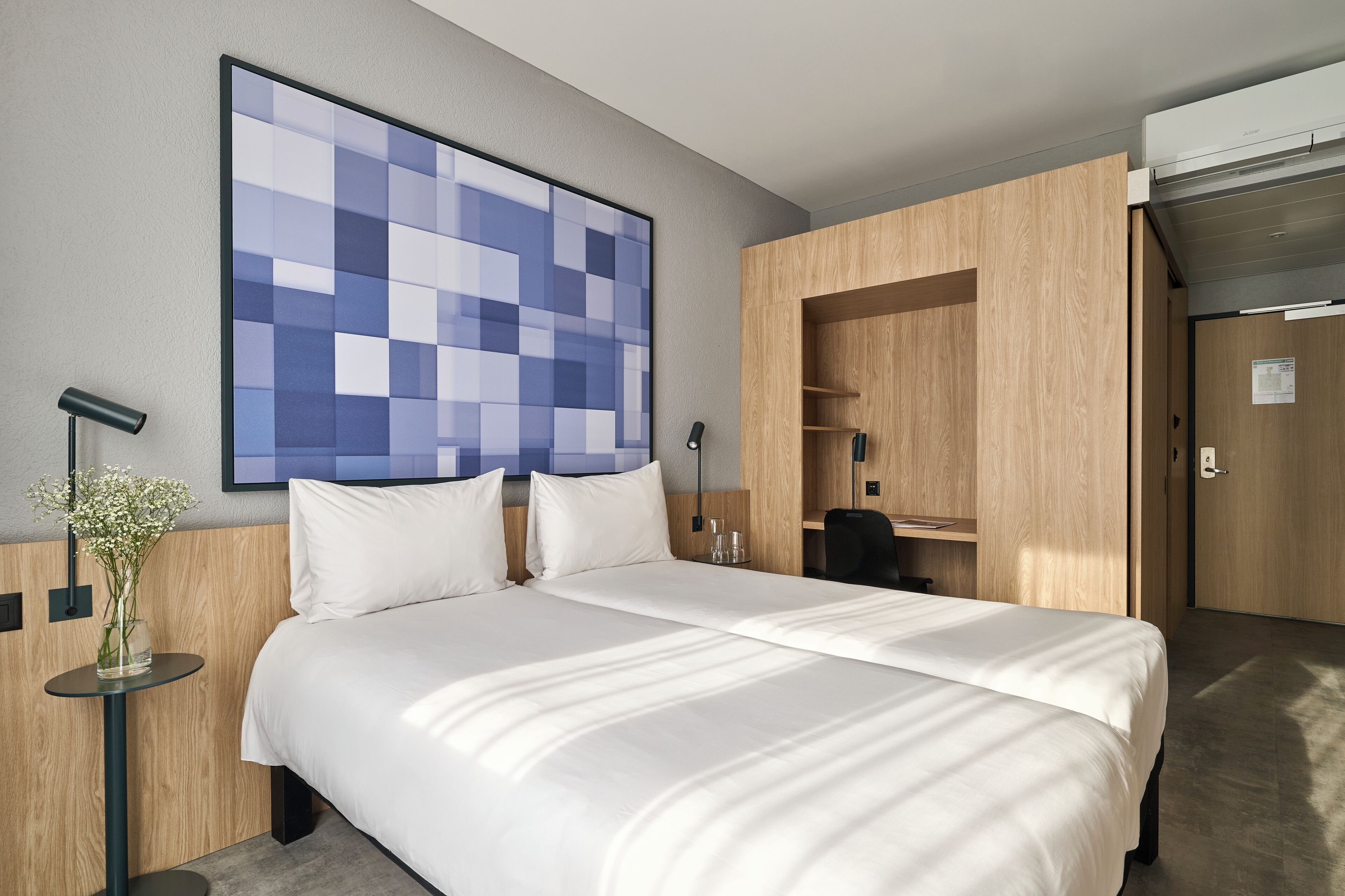 Une chambre d’hôtel avec un grand lit, un téléviseur fixé au mur, une armoire en bois et une petite table sur laquelle est posé un vase.