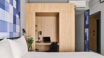 Habitación de hotel con una cama, un escritorio con una computadora portátil, una silla, un florero con flores y un aire acondicionado instalado en la pared.