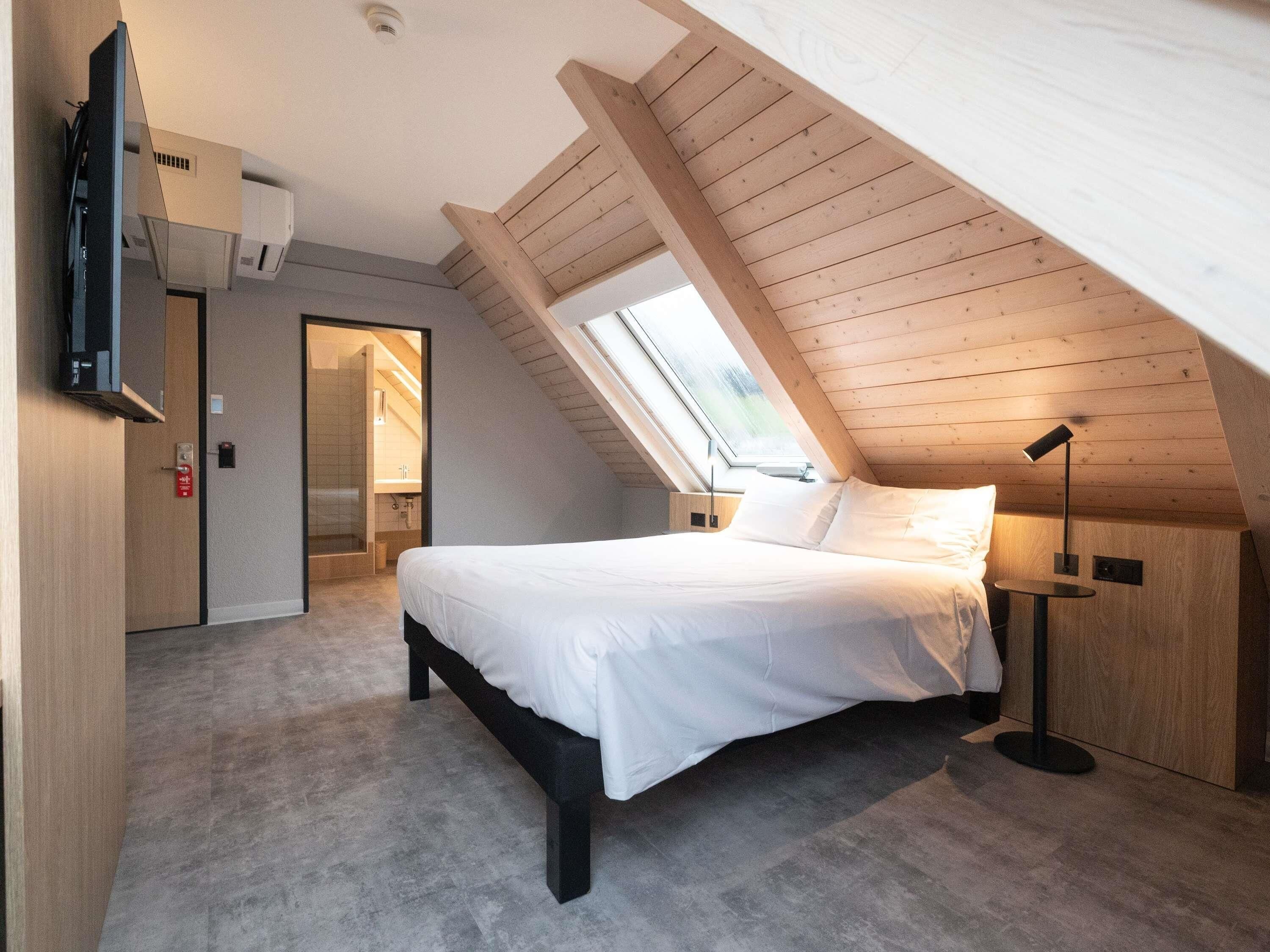 Een slaapkamer met een schuine plafond, een bed, nachtkastje, lamp en een televisie die aan de muur is bevestigd.
