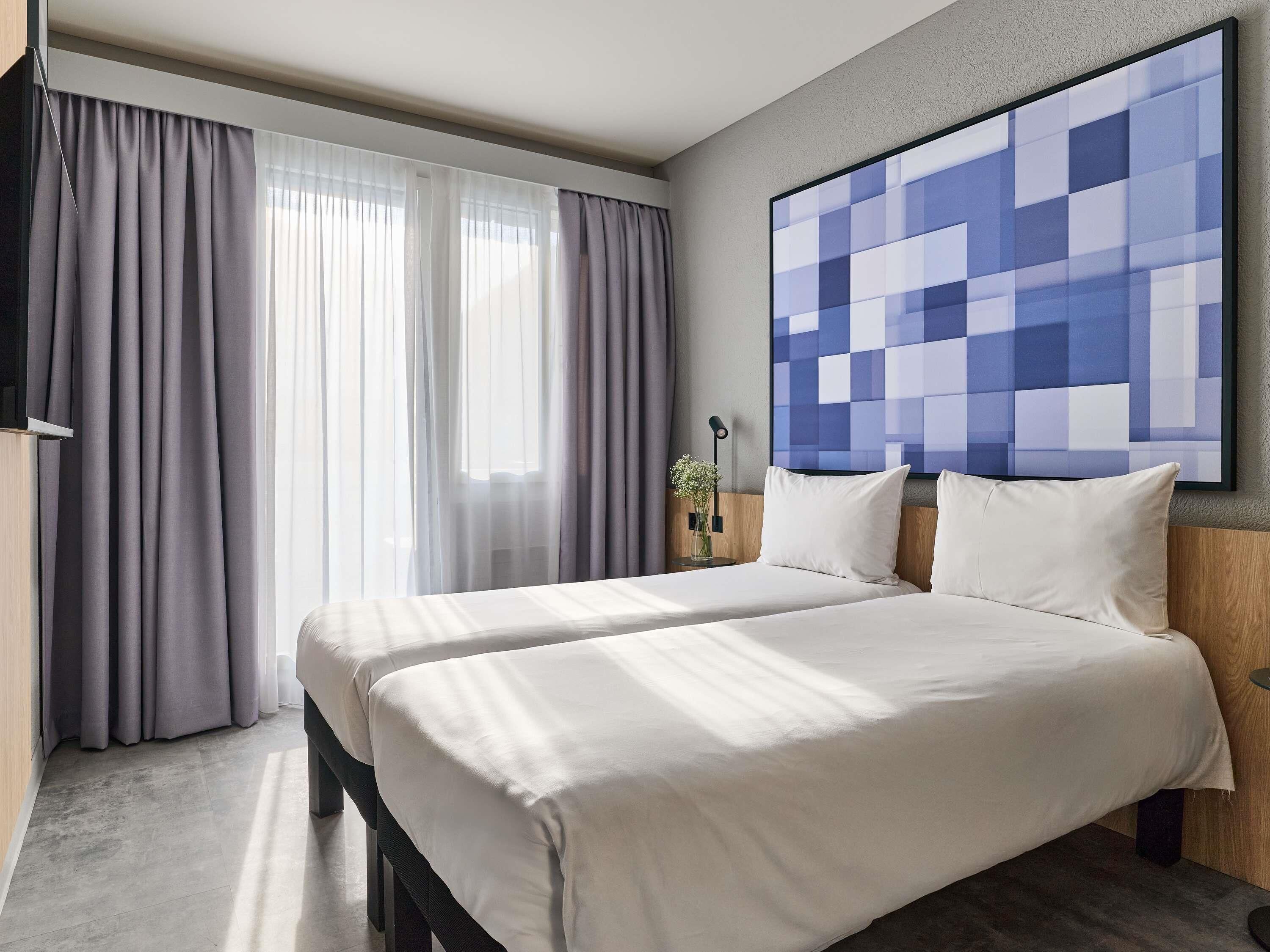 A hotel room with a bed, curtains, a wall-mounted artwork, and a bedside table with a lamp.