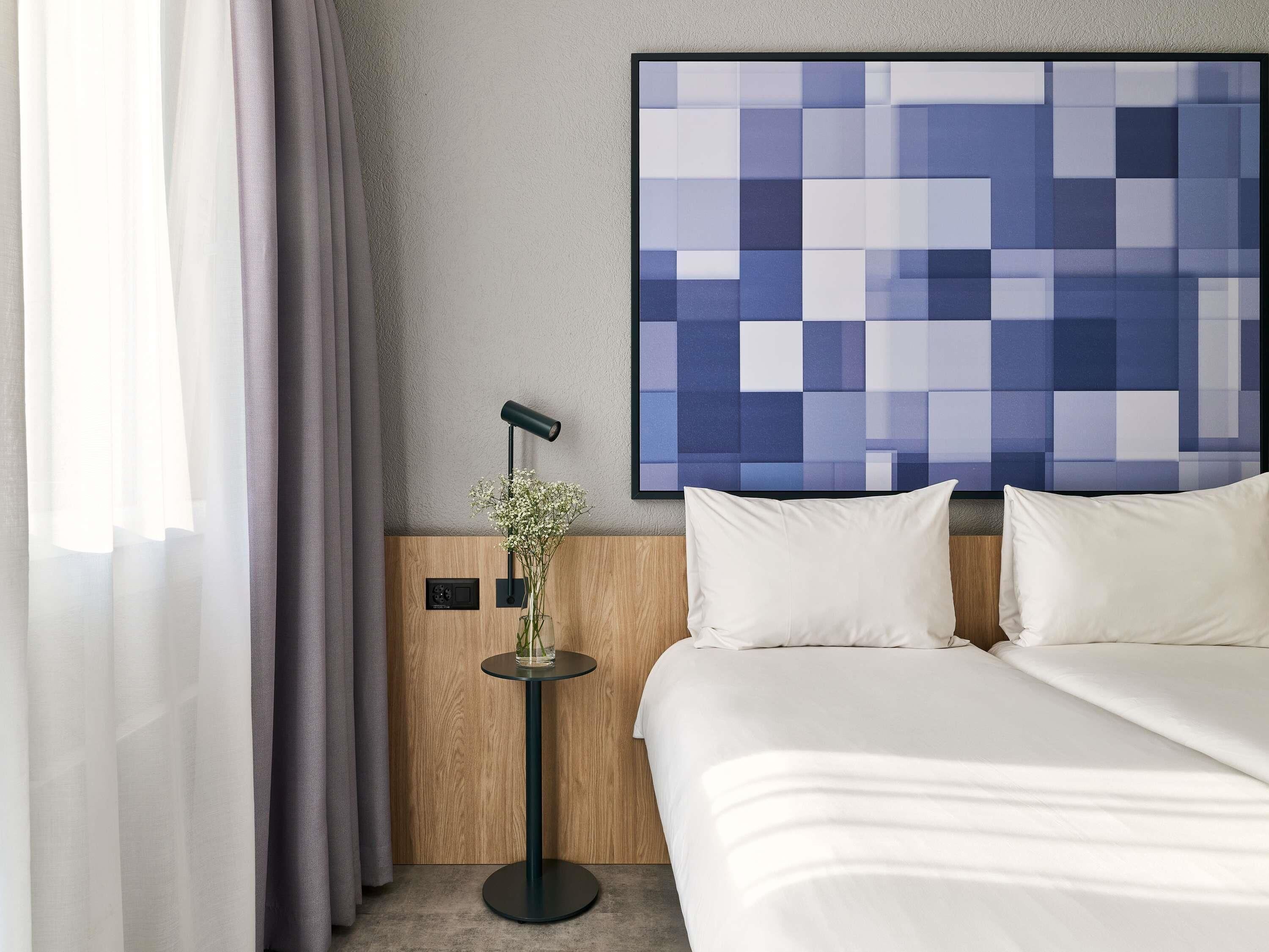 Ein Hotelzimmer mit einem Bett, einem Wandbild, einem Nachttisch mit Lampe und einem Fenster mit Vorhängen.