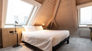 Une chambre moderne avec un plafond en pente, un grand lit, une lampe de chevet et une vue sur le paysage urbain par la fenêtre.