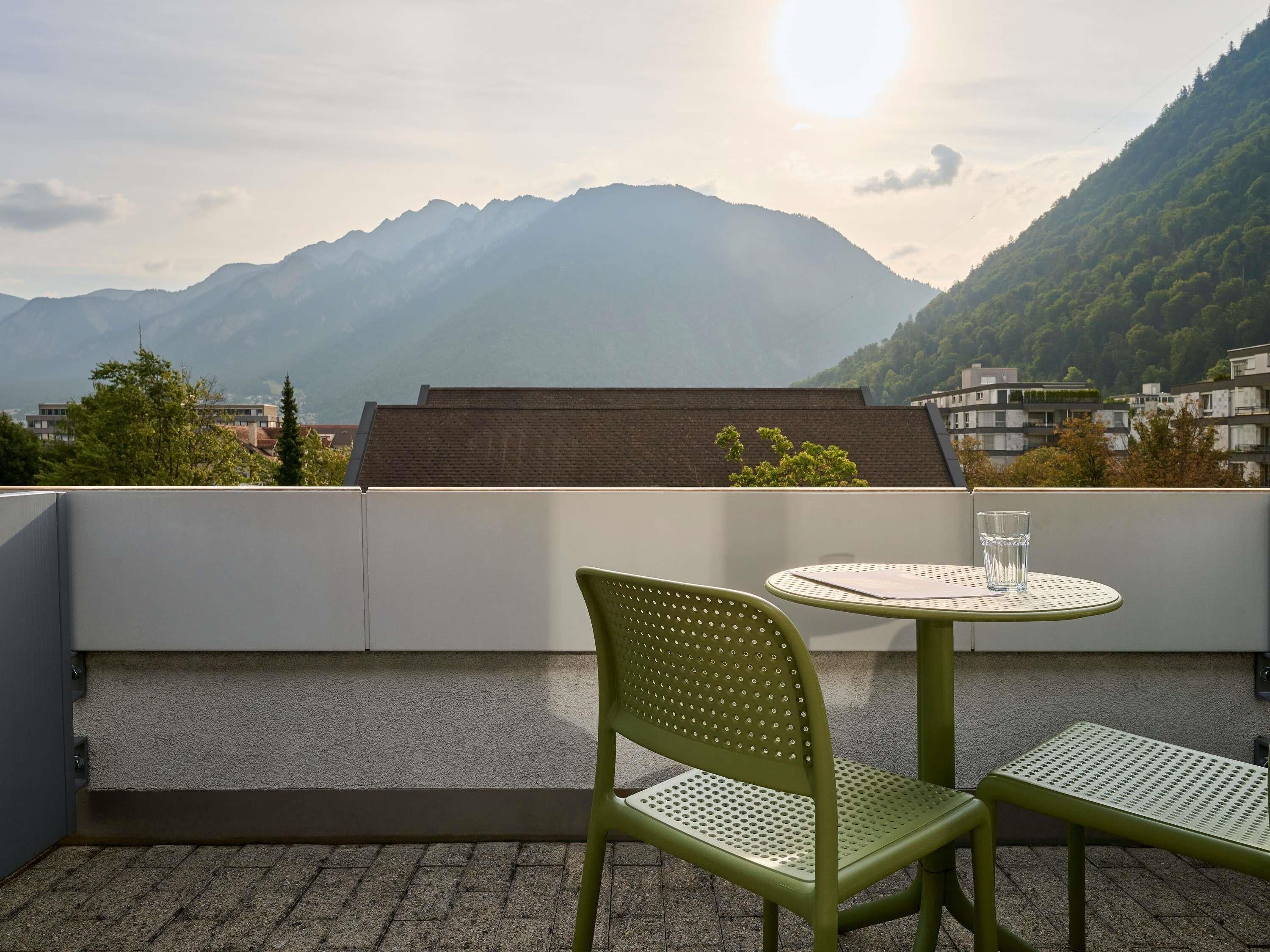 A balcony with a green chair and a small round table, overlooking a mountain and a residential area.