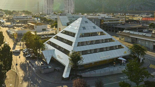 Un restaurant McDonald's doté d'un toit en forme de pyramide, situé au milieu d'immeubles urbains et avec une chaîne de montagnes en toile de fond.