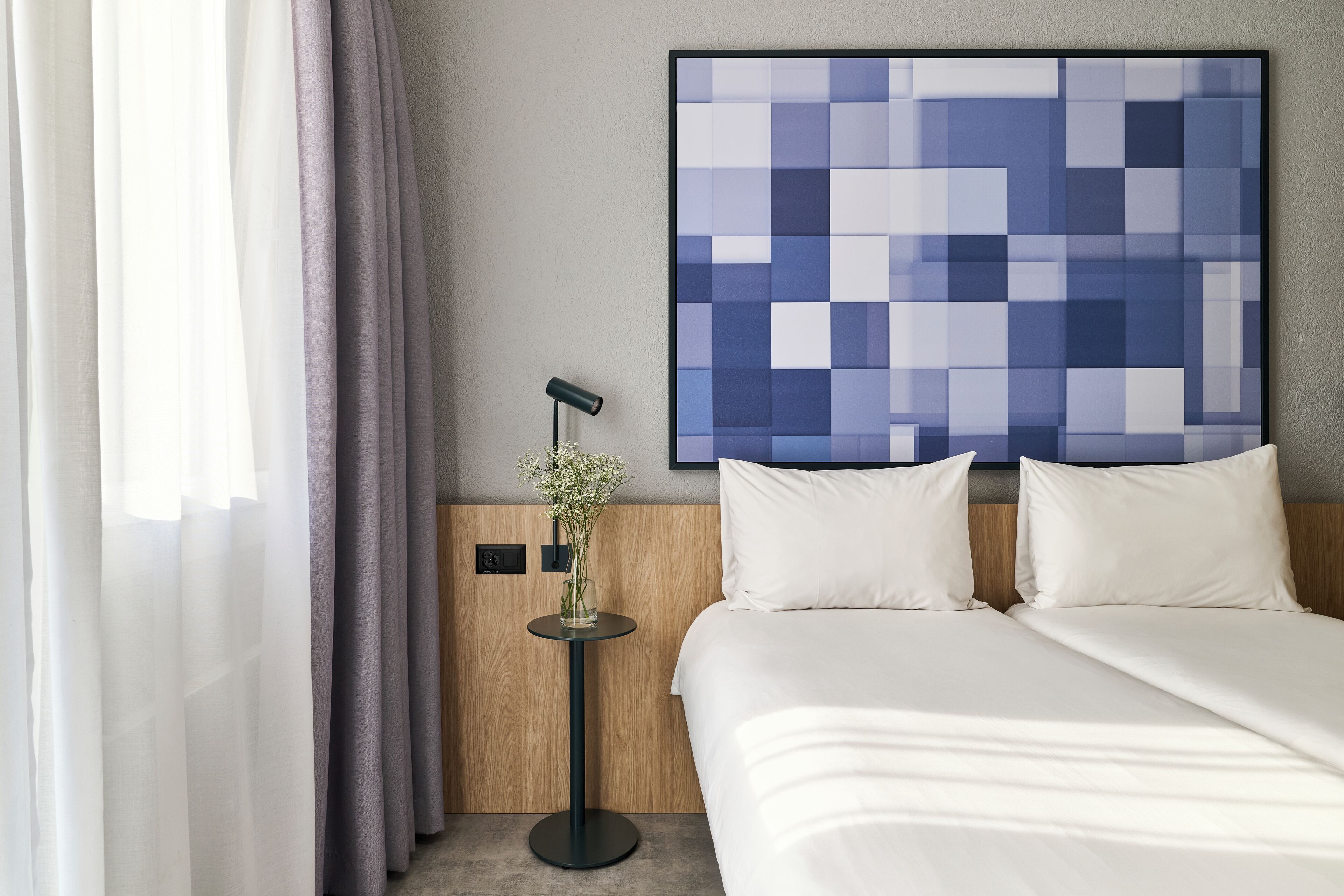Habitación de hotel con cama, ropa de cama blanca, una obra de arte colgada en la pared, una mesita con un florero y una ventana con cortinas traslúcidas.