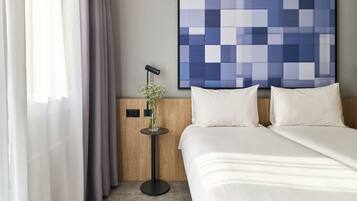 Habitación de hotel con cama, ropa de cama blanca, una obra de arte colgada en la pared, una mesita con un florero y una ventana con cortinas traslúcidas.