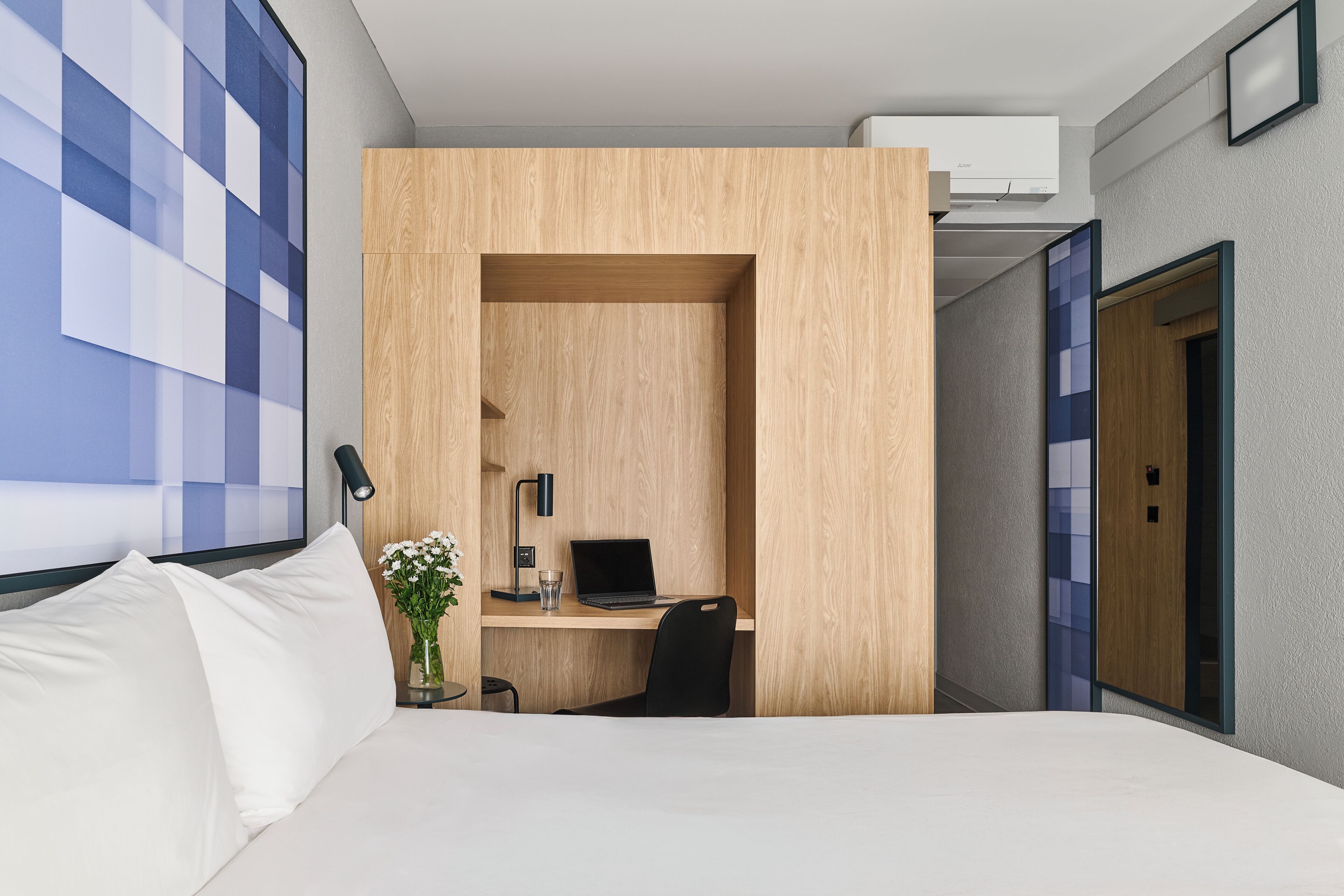 Habitación de hotel con cama, escritorio con una computadora portátil, una silla, un florero con flores y un aire acondicionado instalado en la pared.