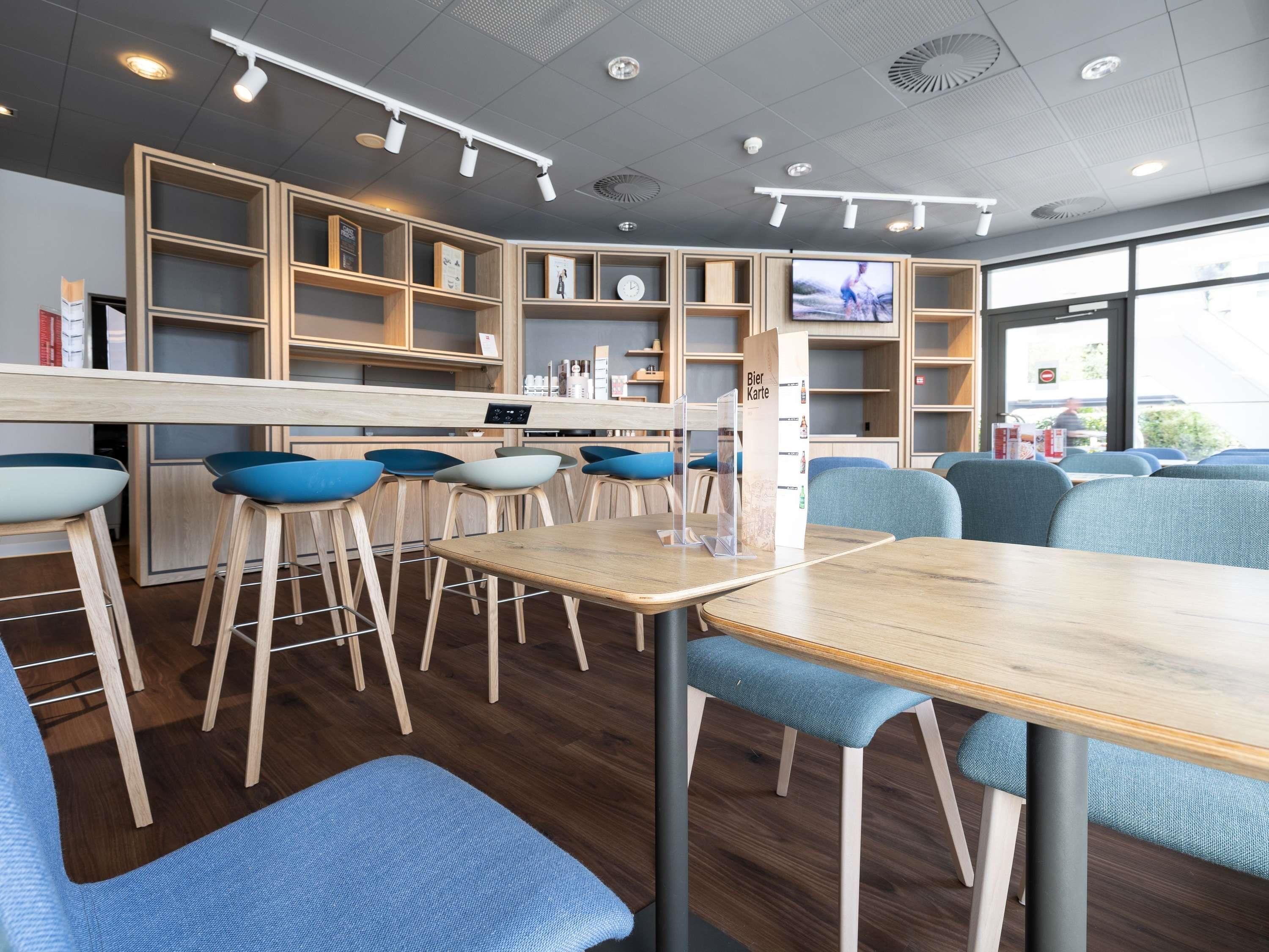 Un café moderno con mesas de madera y sillas azules, una barra con una vitrina de menú, y un ventanal grande que proporciona luz natural.
