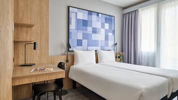 Una habitación de hotel con un escritorio de madera, dos camas, un ventanal con cortinas y una obra de arte colgada en la pared.