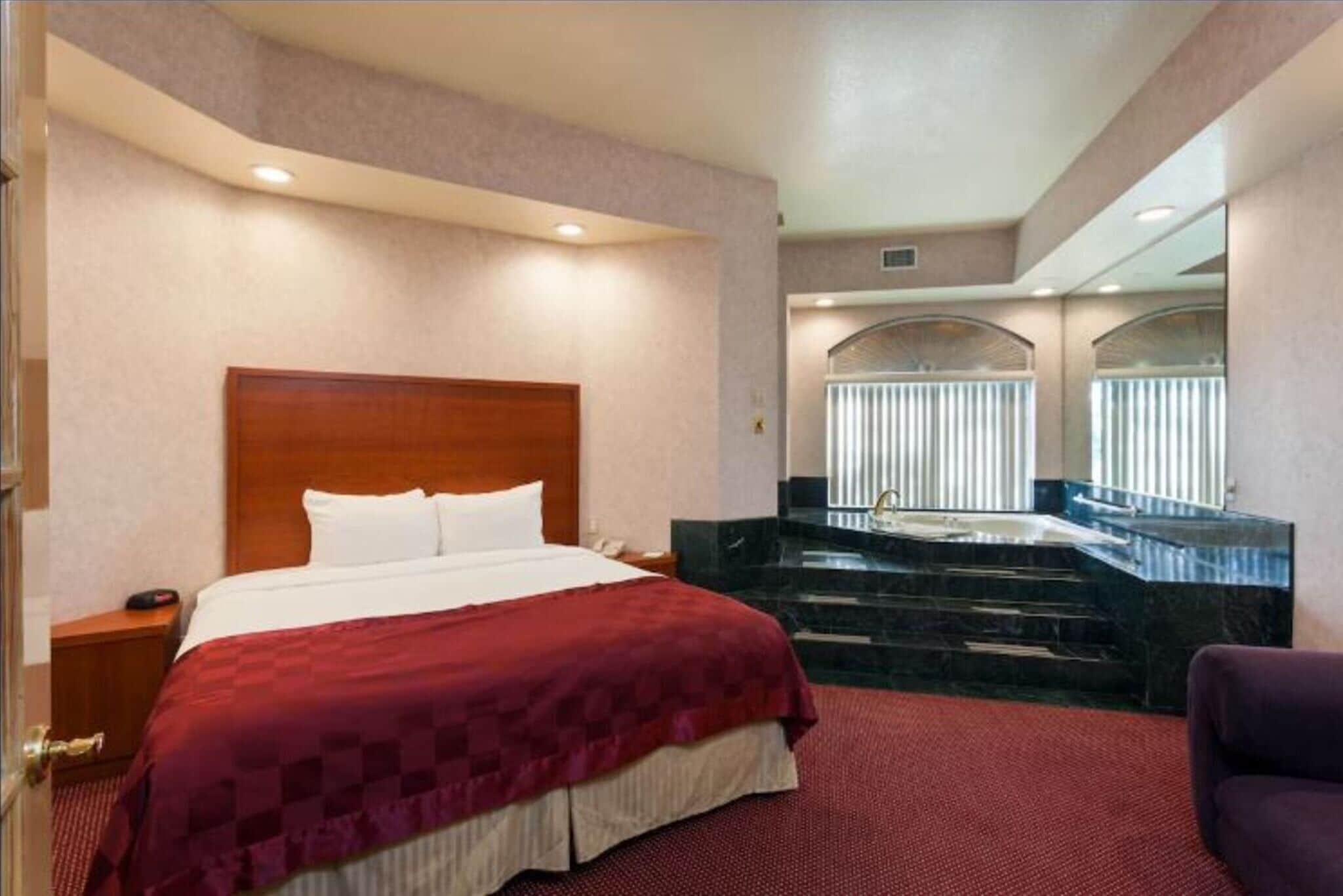 A hotel room with a large bed, a red carpet, a marble bathtub, and a view of the city through the windows.