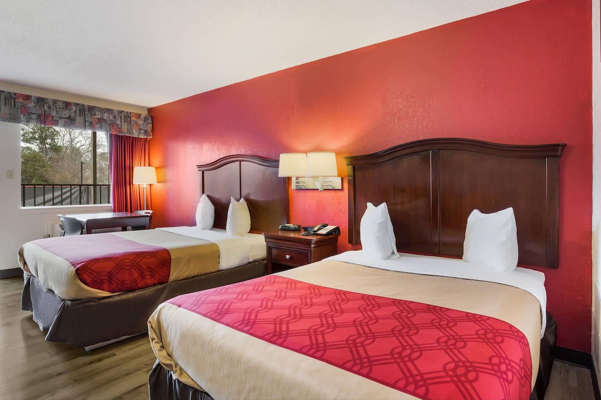 A hotel room with two beds, a desk, a chair, a window with curtains, and a nightstand with a lamp.