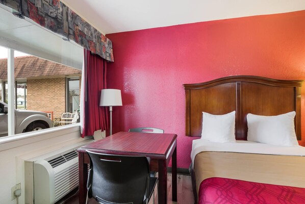 A hotel room with a bed, a wooden headboard, a table with a chair, a window with a view of a car and a building, and a red wall. - Econo Lodge Williamsburg Historic Area (Williamsburg)