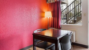 Una habitació d'hotel amb una taula i cadires de fusta, una paret vermella amb textura, una làmpada, una finestra amb cortines i una escala.