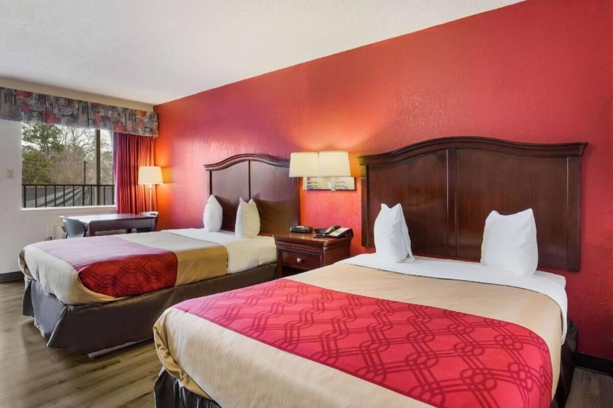 A hotel room with two beds, a desk, a chair, a window with curtains, and a nightstand with a lamp.