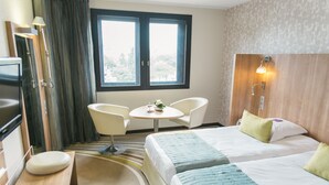 Une chambre d’hôtel avec deux lits, une petite table ronde, deux chaises, un bureau avec une chaise et une fenêtre avec des rideaux.