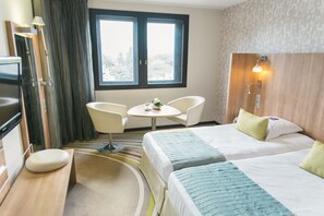 A hotel room with two beds, a small round table, two chairs, a desk with a chair, and a window with curtains.