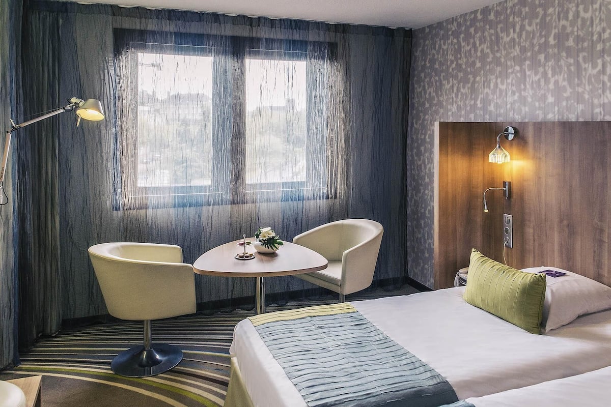 A hotel room with a bed, two chairs, a small table, a lamp, and a window with curtains.