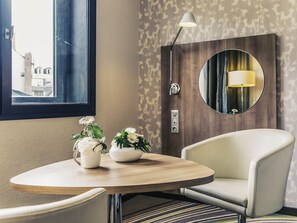 A hotel room with a round wooden table, two white flower vases, a modern armchair, a wall-mounted lamp, and a mirror reflecting a lamp. - Mercure Angers Centre De Congres (Angers)