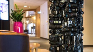Une exposition d’appareils photo disposés en une pile verticale haute, sur un socle noir, dans un hall d’entrée.