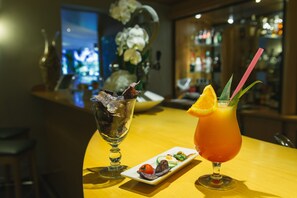 A bar setting with a cocktail and a glass of ice cubes, a small plate of appetizers, and a vase of flowers in the background.
