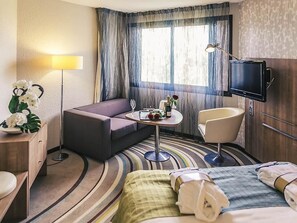A hotel room with a bed, a sofa, a small table with a vase and flowers, a chair, a TV, and a desk with a lamp.