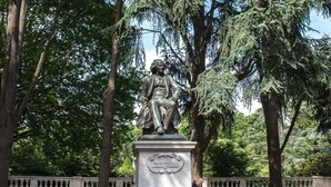 Une statue représentant deux personnages assis sur un piédestal, dans un parc avec des arbres et une clôture en arrière-plan.