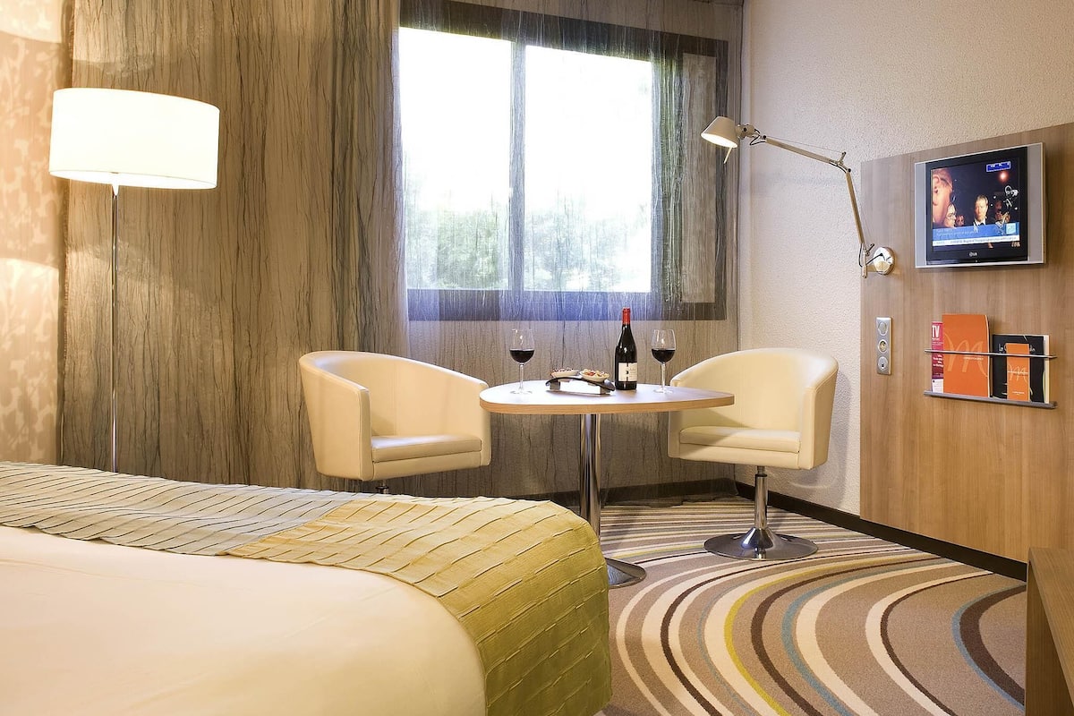 A hotel room with a bed, two chairs, a small table with wine, a TV, and a window with curtains.