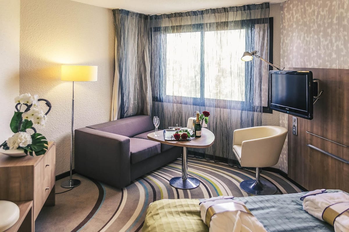 A hotel room with a sofa, a small table with a bottle and flowers, a chair, a bed, a TV, and a window with curtains.