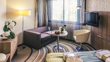 Habitación de hotel con sofá, una mesita con una botella y vasos, una silla, cama con toallas, televisión y ventana con cortinas.