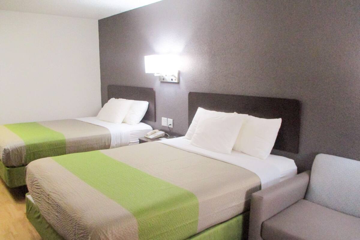 A hotel room with two beds, a nightstand, a lamp, and a chair.