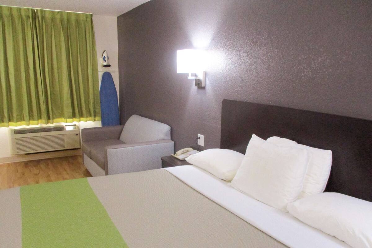 A hotel room with a bed, a chair, a bedside table with a phone, and a wall-mounted light.