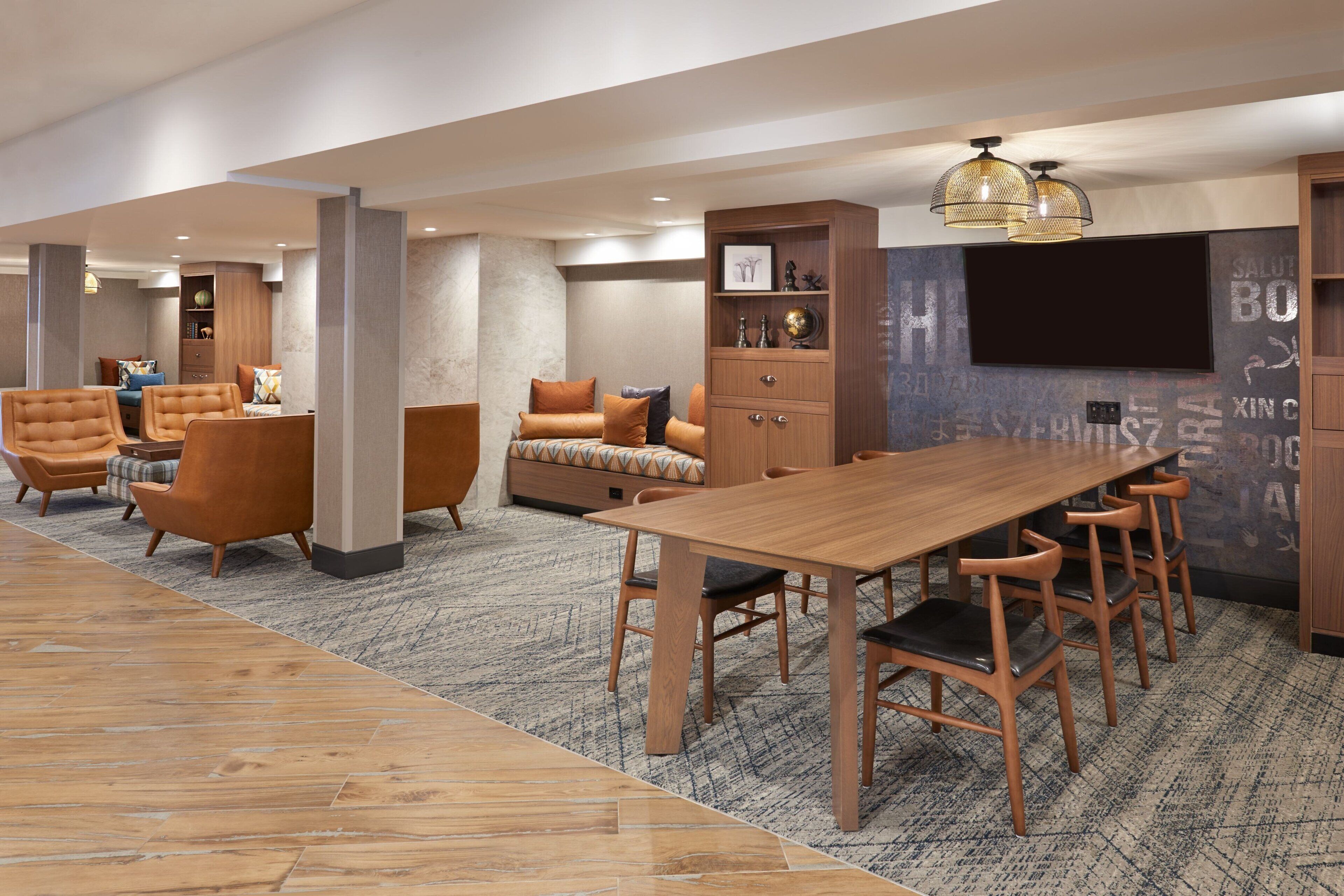 Un hall d’hôtel moderne avec une table à manger en bois, des sièges confortables, un téléviseur à écran plat et un mur décoratif orné de motifs textuels.