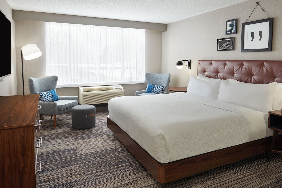A hotel room with a large bed, a desk, a chair, a lamp, and a window with blinds.