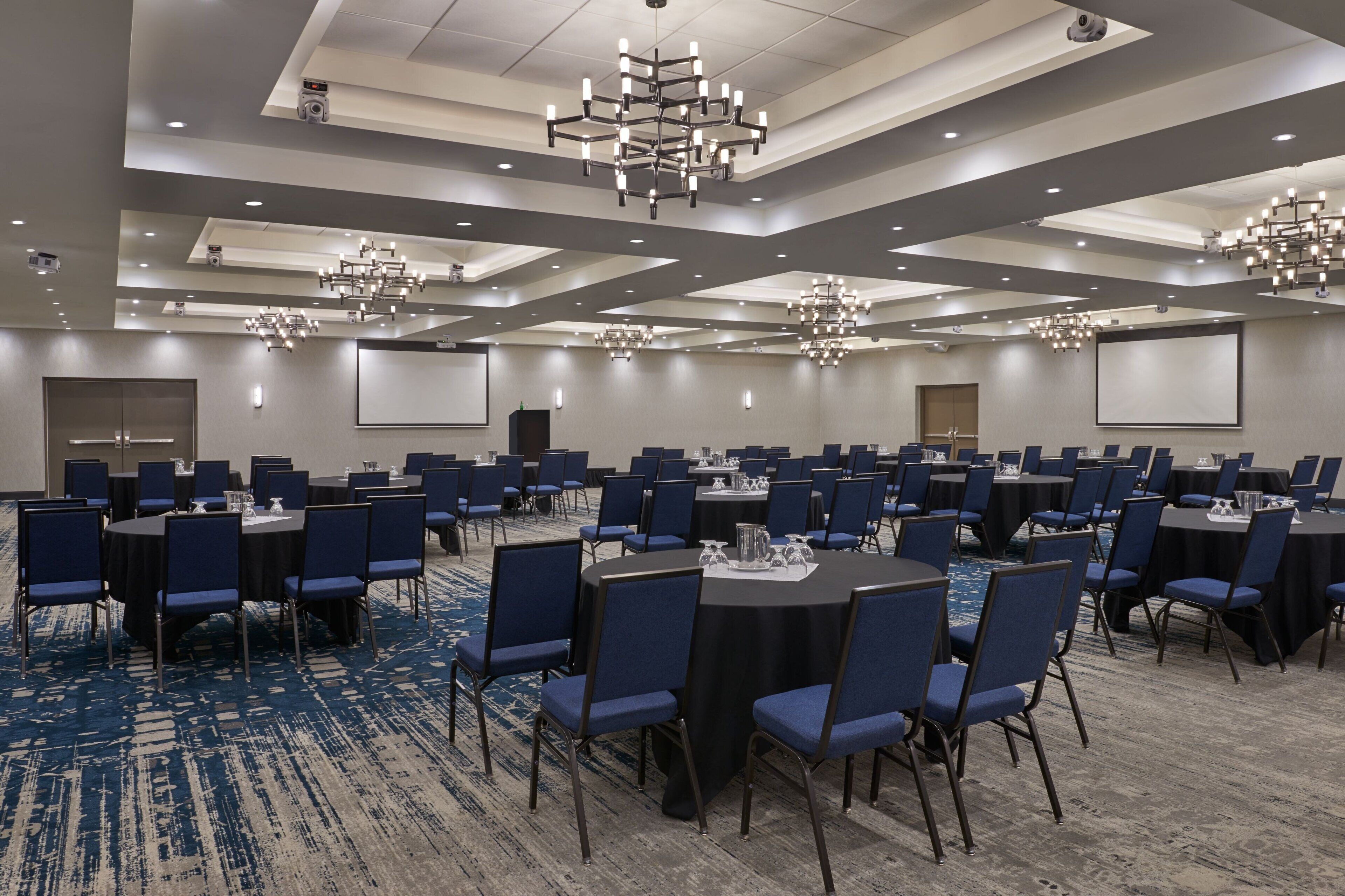 A large conference room with multiple tables and chairs, equipped with projectors and screens, and a modern chandelier.