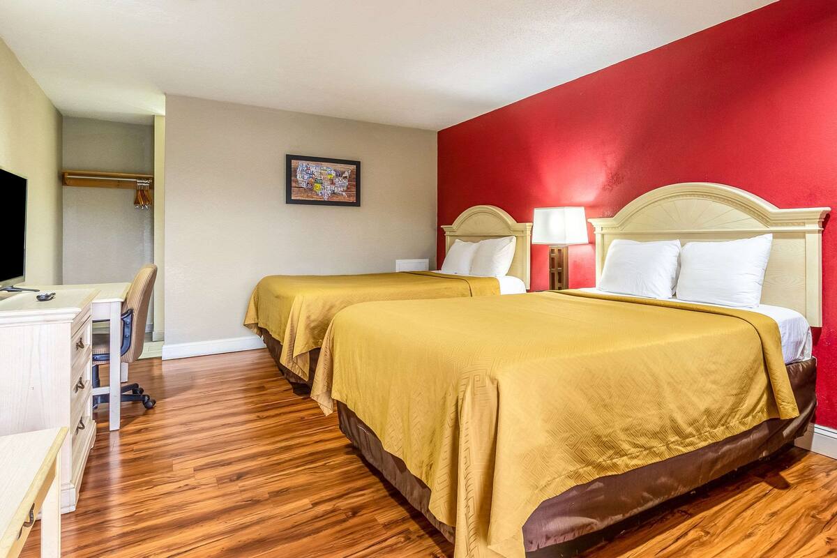 A hotel room with two beds, wooden flooring, a desk, and a red accent wall.