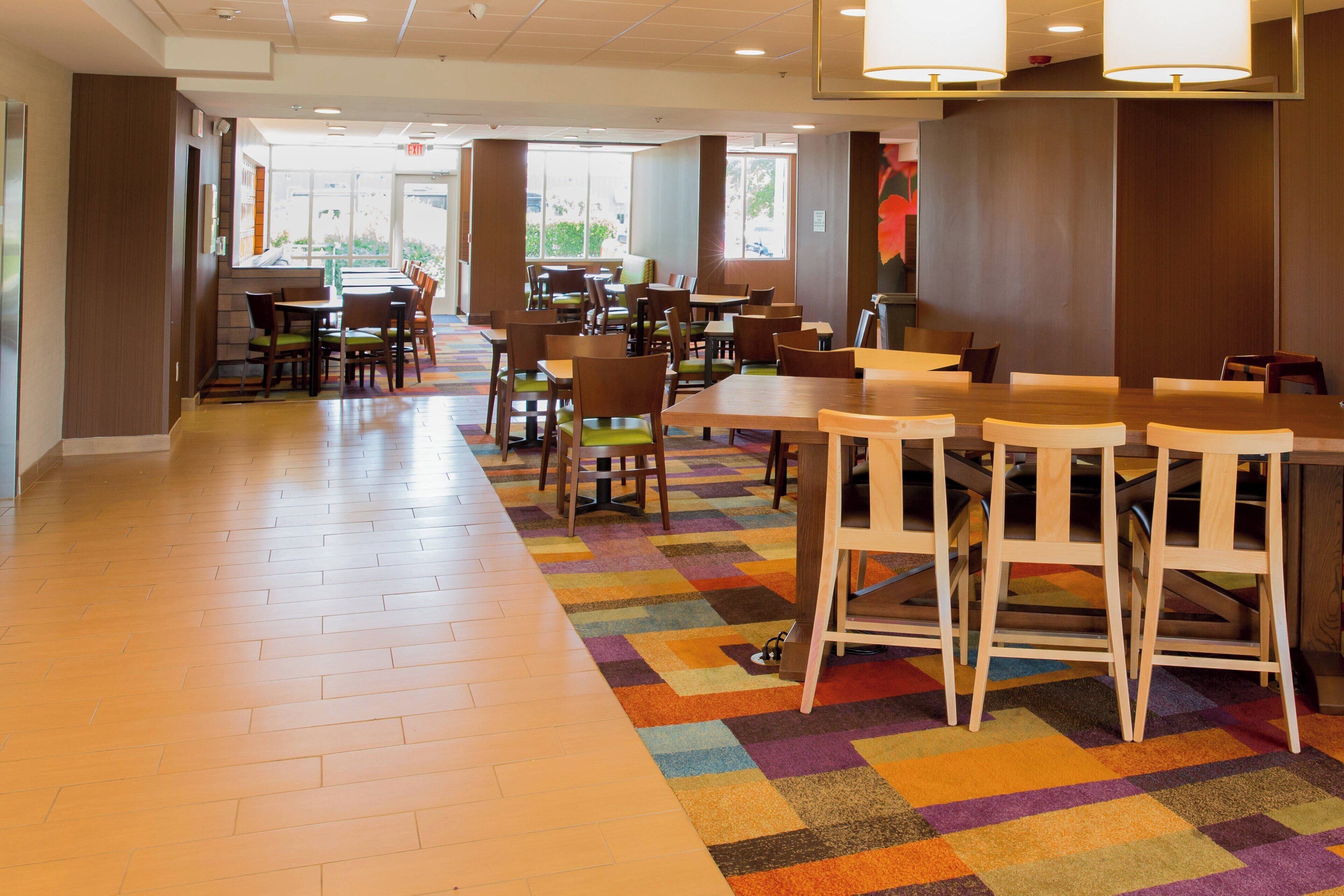 Una amplia cafetería con numerosas mesas y sillas, una alfombra colorida y grandes ventanales que dejan entrar luz natural.
